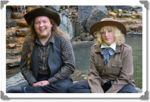 Noah and Acacia are wearing costumes and are sitting in front of a natural pool. Noah is wearing a cowboy hat.