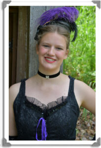 A headshot of Freya in costume. She is wearing a cancan dress with a purple feather fascinator.